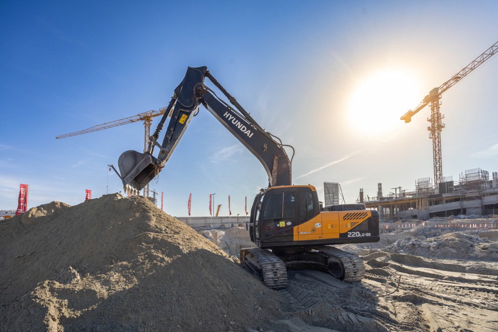 Cotier House 2 By Imtiaz A Hyundai excavator operates on a construction site, lifting sand under a clear sky with cranes in the background.